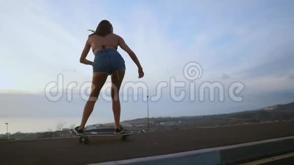 穿短裤和运动鞋的女孩在海洋和山脉的背景下沿着道路滑板视频的预览图
