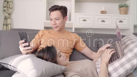 亚洲夫妇在家里的客厅里使用智能手机和笔记本电脑检查社交媒体甜蜜的夫妇享受爱情的时刻视频的预览图
