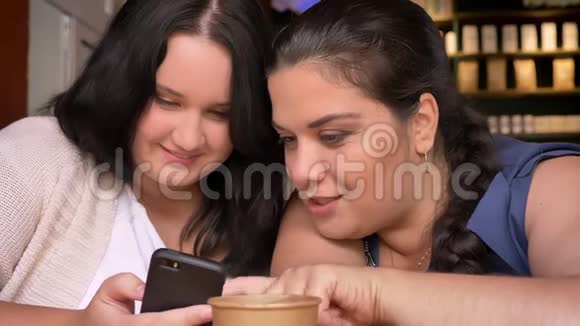两个肥胖的白种人年轻模特一边在咖啡馆里拿着纸杯一边偷着智能手机视频的预览图