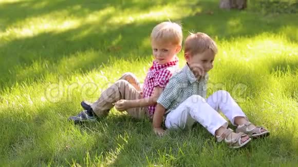 两个4岁的小男孩背靠背坐在草地上的公园里视频的预览图