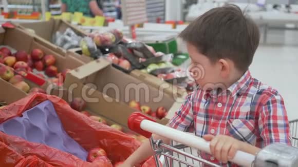 有趣的小男孩在超市的家庭购物中坐在手推车里的盒子里摘苹果视频的预览图