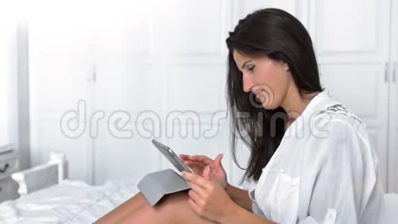 中镜头微笑的幸福女人坐在卧室的床上用平板电脑看屏幕视频的预览图