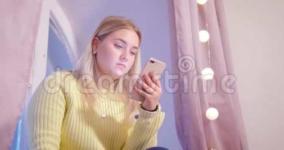 一个丰满的金发女孩坐在她的房间里背景是粉红色的窗帘看着手机屏幕然后她是视频的预览图