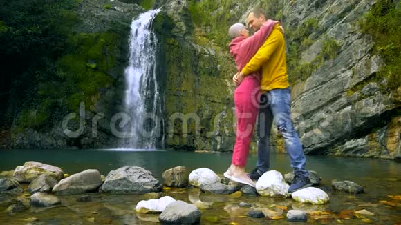 夫妇一个年轻人和一个秃头女孩在瀑布的背景上拥抱和亲吻旅行和家庭度假概念视频的预览图