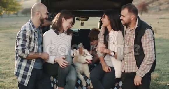 特写两家人带着一只小哈士奇狗和一个小男孩坐在车的后备箱里玩得很开心视频的预览图