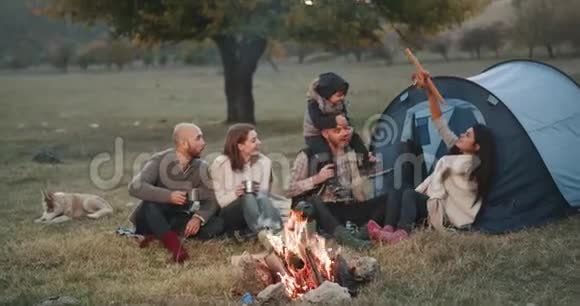 美丽的年轻家庭和他们的朋友在野餐时坐在篝火旁爸爸和妈妈在玩木头视频的预览图