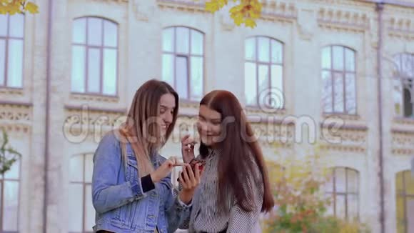 在大学附近的秋天公园里两个美丽的微笑女孩正在看电话视频的预览图