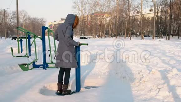 穿着蓝羽绒服带引擎盖的女人在冬季公园侧景中做滚动模拟器练习视频的预览图