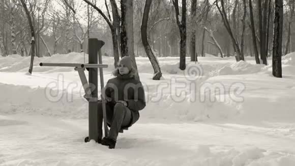 穿蓝色羽绒服带头罩的人在冬季公园前景BW模拟器上做腿部运动视频的预览图