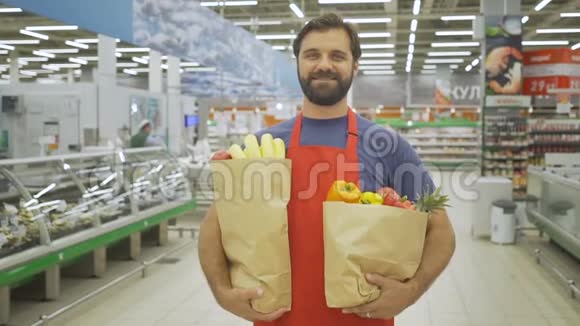 英俊的售货员拿着购物袋站在超市他正对着照相机微笑视频的预览图
