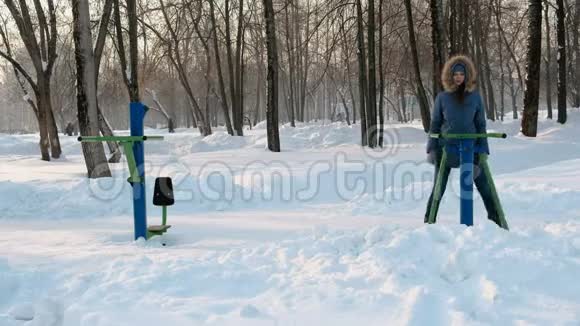 穿着蓝色夹克的女人正在模拟器上做腿部运动在冬季公园的正面观看视频视频的预览图