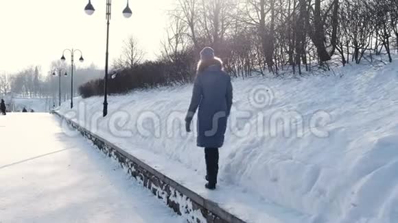 穿着蓝色羽绒服的年轻女子在冬季公园沿着边境散步看着天空回头看视频的预览图