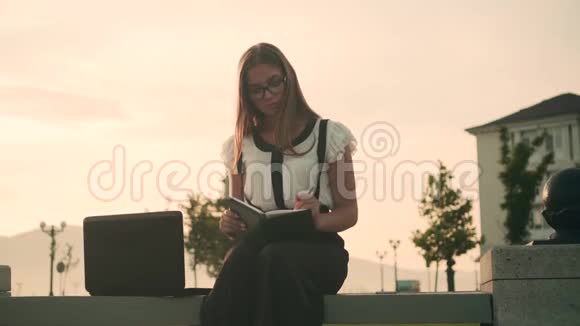 商务女性穿着白色衬衫和眼镜坐在长凳上工作女孩写在笔记本里看着笔记本电脑视频的预览图