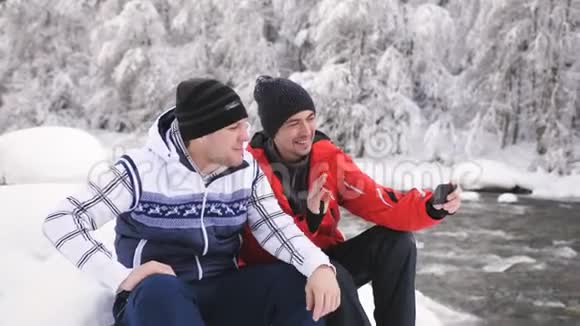 冬天两个开朗的男人在河边与朋友用视频电话在智能手机上交流视频的预览图