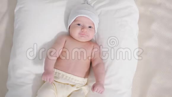 从上面拍摄的可爱的婴儿戴着帽子和裤子躺在灰色背景下看着相机视频的预览图