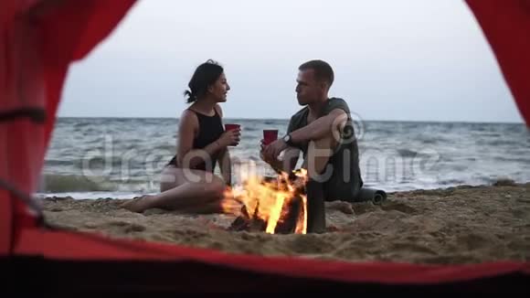 年轻的夫妇男人和女人一起在靠近火的海滩上喝着红色的乳状杯录像视频的预览图