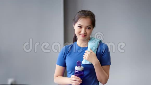 肖像微笑亚洲女健身教练拿着瓶水和毛巾看着相机视频的预览图