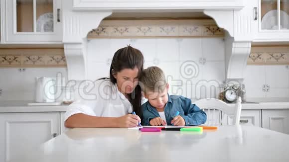 年轻漂亮的妈妈在家里的厨房里用彩色毡尖笔教她的小儿子画画视频的预览图