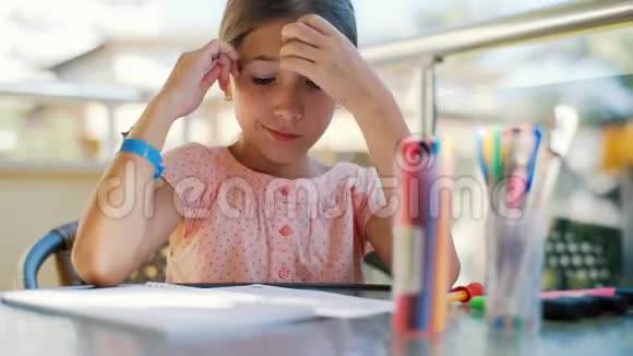 可爱的女孩在笔记本上画一支铅笔坐在阳台上的玻璃桌上女孩在笔记本上画画唱着视频的预览图