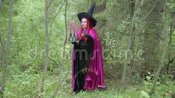 年轻的女巫红发黑帽子覆盆子斗篷在森林里点燃蜡烛视频的预览图