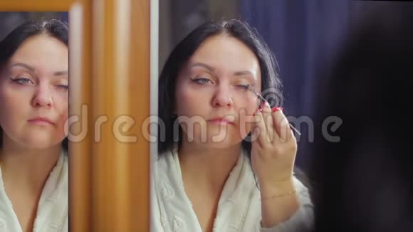 一个穿着白大褂的年轻黑发女人在镜子前用刷子把化妆品的阴影涂在她的眼皮上视频的预览图