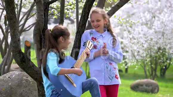 快乐和积极的两个学校女朋友在自然公园玩音乐和唱歌一个年轻女孩弹吉他另一个女孩拍手视频的预览图