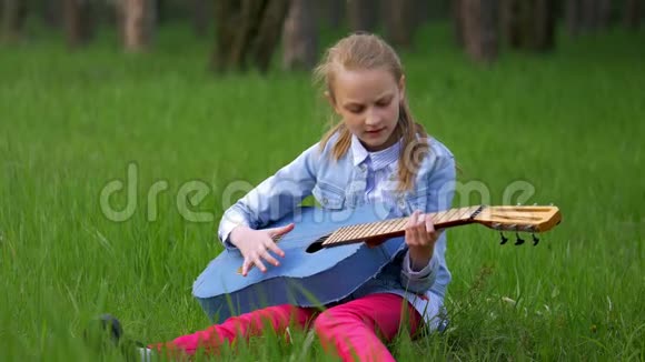 穿着牛仔裤夹克的小女孩玩得很开心在春天坐在绿草如茵的森林里弹吉他视频的预览图