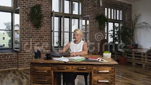 漂亮的中年白种人女士戴着眼镜在上班期间坐在桌旁带着笔记本电脑在时尚的办公室里视频的预览图