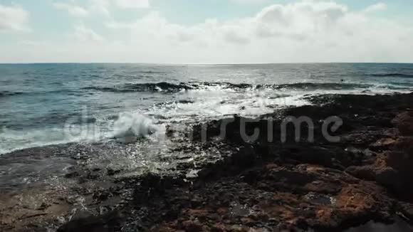 空中拍摄的海浪击中岩石海滩溅到岩石上蓝天地平线上有太阳之路多风的da海洋视频的预览图