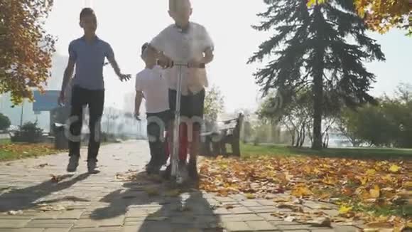 这个男孩在秋天的公园里骑滑板车朋友们正在追赶一个在户外骑滑板车的男孩孩子们玩游戏视频的预览图
