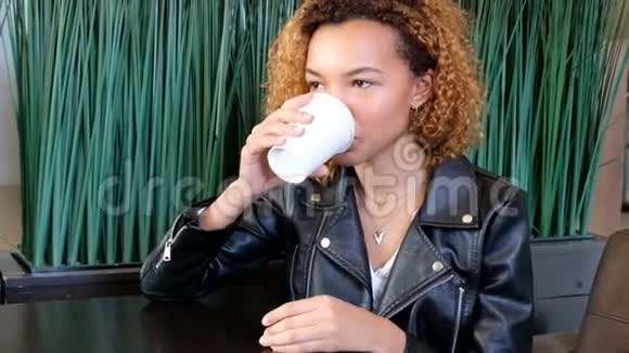 一个身穿皮夹克的年轻黑皮肤漂亮的女孩正在绿色的咖啡馆里喝着一杯白色的咖啡视频的预览图