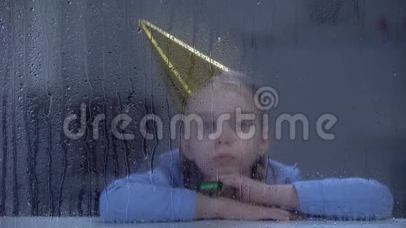 孤独的小女孩戴着生日帽和派对鼓风机看着雨窗视频的预览图