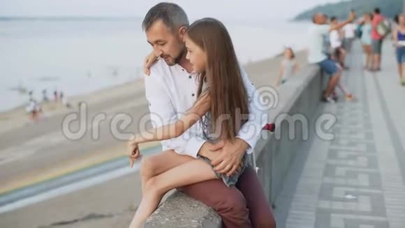 一个女孩坐在她父亲腿上一男一女坐在堤上日落4k视频的预览图