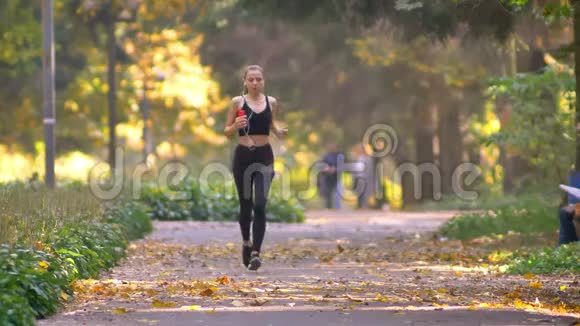 穿着连衣裙的年轻女孩带着耳机在秋季公园跑步停下来系紧她的短裤继续慢跑视频的预览图