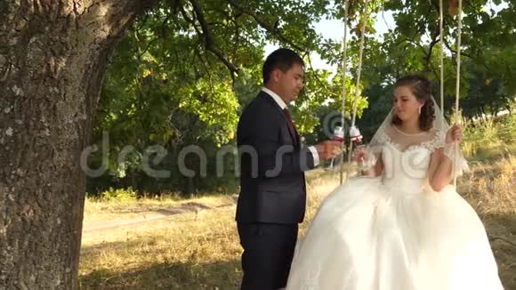 快乐的新郎和穿白衣服的新娘从漂亮的酒杯里喝香槟在公园里荡秋千视频的预览图