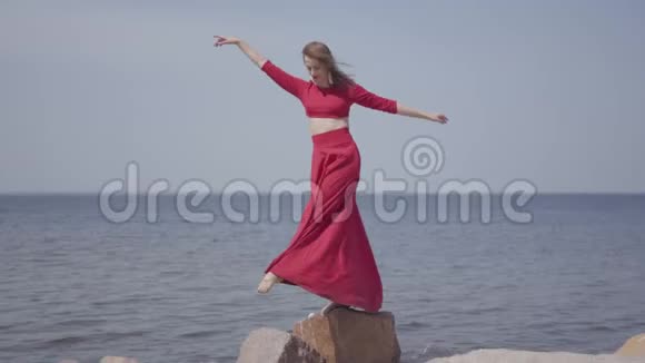 穿着红色长裙跳舞的可爱的年轻女人在蓝色海景附近的大石头上举手视频的预览图