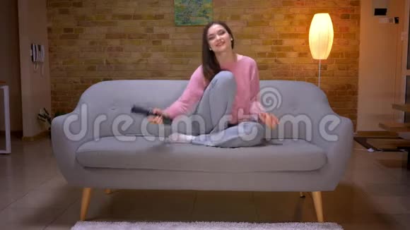 一位年轻漂亮的黑发白种人女性坐在沙发上兴奋而好奇地看电视的特写镜头视频的预览图