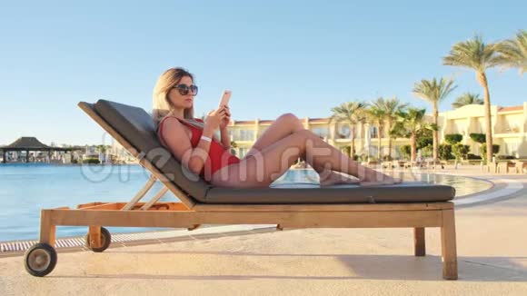 漂亮的年轻女士穿着红色比基尼和太阳镜躺在甲板椅上使用智能手机应用程序并在酒店发短信视频的预览图