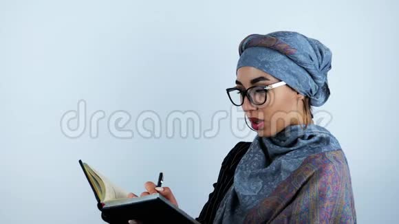 年轻漂亮的阿拉伯商务女性戴着眼镜翻着笔记本手里拿着笔白色的背景上视频的预览图