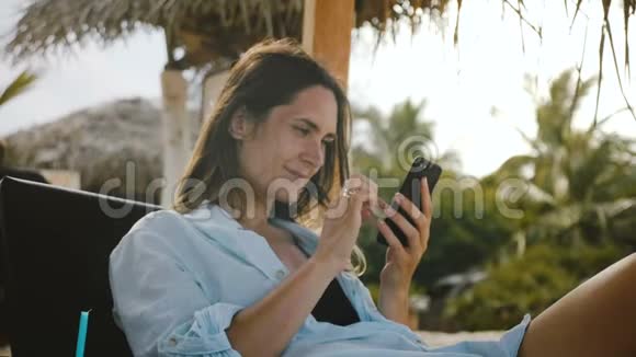 相机放大了快乐美丽的年轻女子使用智能手机应用程序微笑休息在异国情调的海滩度假椅视频的预览图