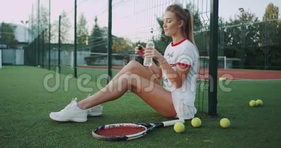 喝着一瓶水可爱的女士在外面的网球场上坐在地板上晴天视频的预览图