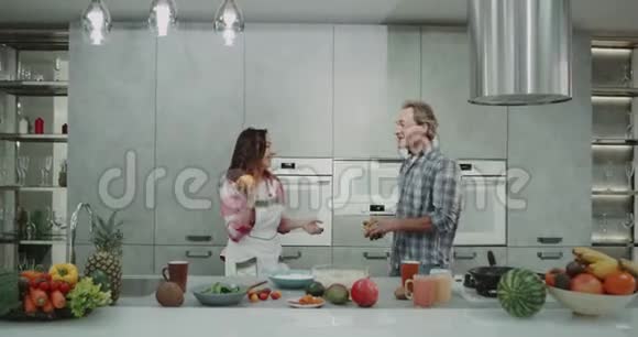 有趣的成熟夫妇在一个现代化的灰色厨房里和橘子一起杂耍同时做早餐4k视频的预览图
