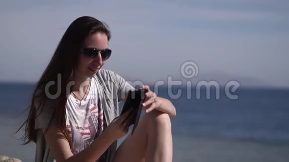 一个年轻女孩戴着太阳镜坐在海滩上打电话视频的预览图