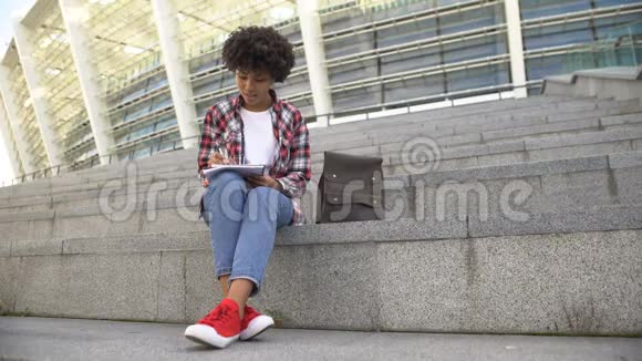 可爱的卷发女学生坐在竞技场附近的楼梯上做作业视频的预览图