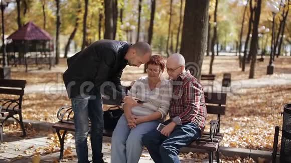 成年儿子在秋季公园的智能手机上显示年迈的父母照片视频的预览图