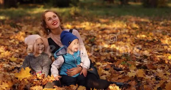 美丽的年轻母亲和两个孩子在树叶上享受阳光明媚的秋日视频的预览图