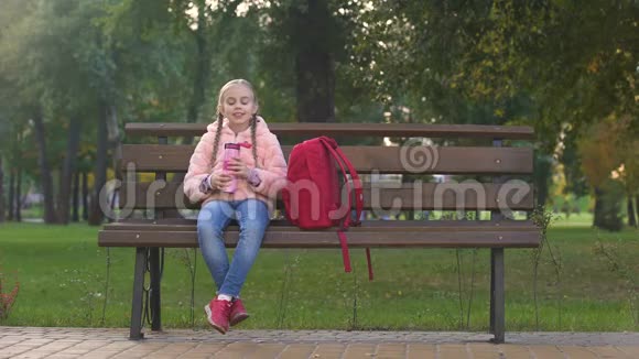 快乐的女学生拿着背包一边坐在长凳上一边从热水杯里喝茶视频的预览图