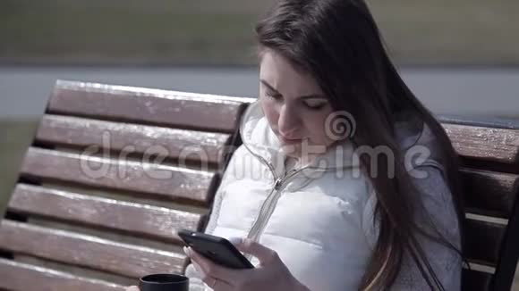 美丽的年轻女孩坐在城市公园的木凳上穿着白色西装喝着热杯咖啡视频的预览图