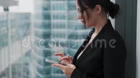 一位女商人站在一座摩天大楼靠近窗户的高楼上用手机视频的预览图