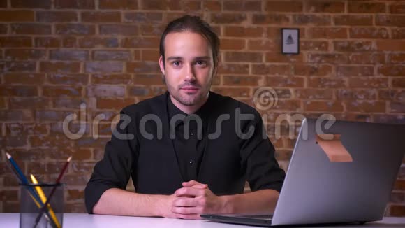 一个英俊的白种人办公室男人一边看着相机一边微笑一边坐在靠近笔记本电脑的办公桌上视频的预览图